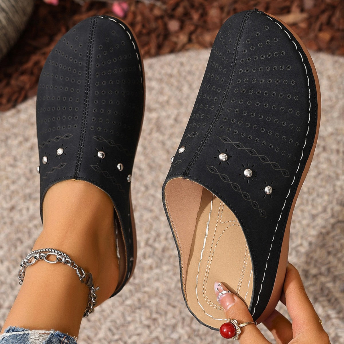 Black slippers with pearl embellishments worn by a person on a textured surface.