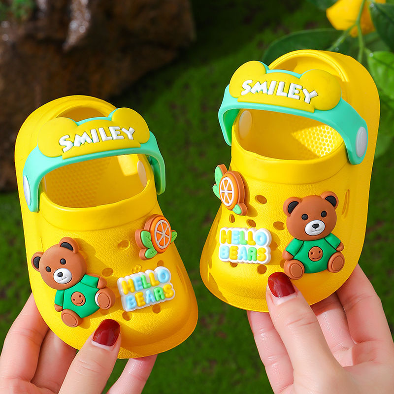 Yellow children's sandals with bear design and 'Smiley' branding held by a person against a green background.